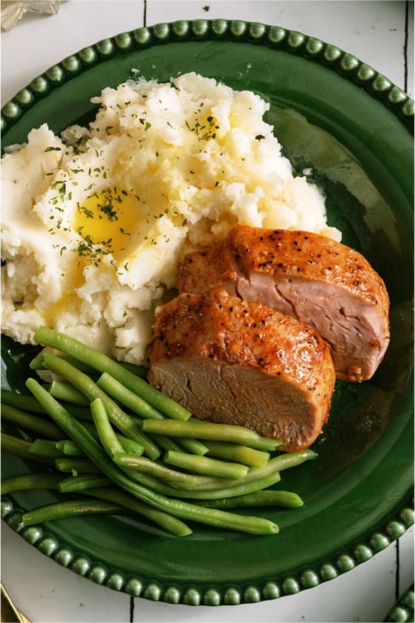 Two slices of Oven Baked Pork Tenderloin on a plate with mashed potatoes and green beans.