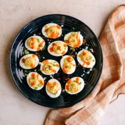 A black plate holds twelve deviled eggs topped with sauce and green garnish, placed next to a folded brown and white checkered cloth on a light countertop.