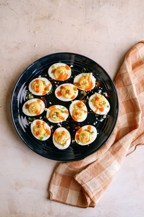 A black plate holds twelve deviled eggs topped with sauce and green garnish, placed next to a folded brown and white checkered cloth on a light countertop.