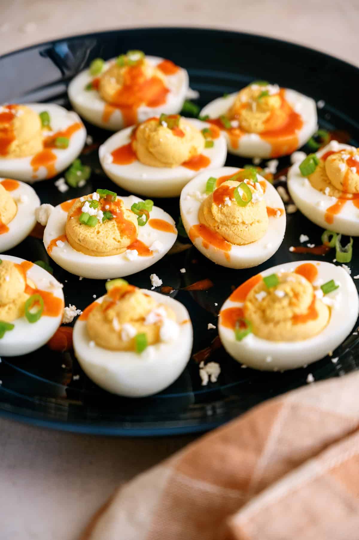 A black plate of deviled eggs garnished with sliced green onions, hot sauce, and crumbled cheese. An orange and white napkin is partially visible in the foreground.