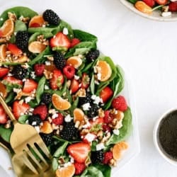 A spinach salad topped with strawberries, raspberries, blackberries, mandarin slices, walnuts, and crumbled cheese is served with dressing on the side.