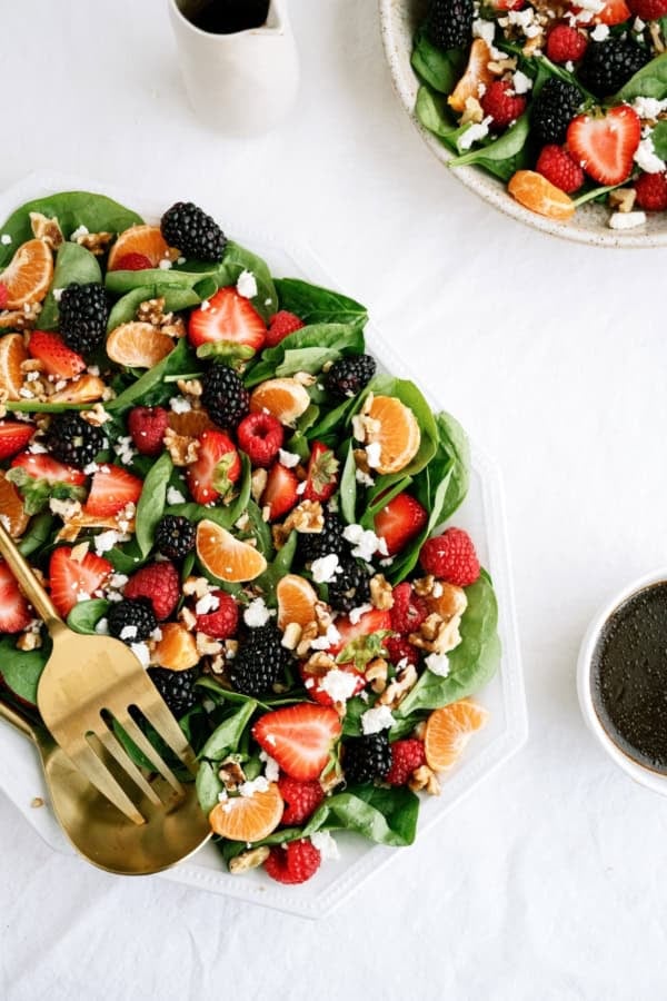 A spinach salad topped with strawberries, raspberries, blackberries, mandarin slices, walnuts, and crumbled cheese is served with dressing on the side.