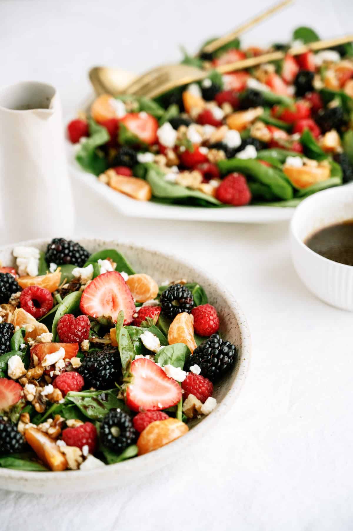 A bowl of spinach salad topped with strawberries, blackberries, raspberries, mandarin slices, nuts, and cheese, with a side of dressing. Another platter and utensils in the background.