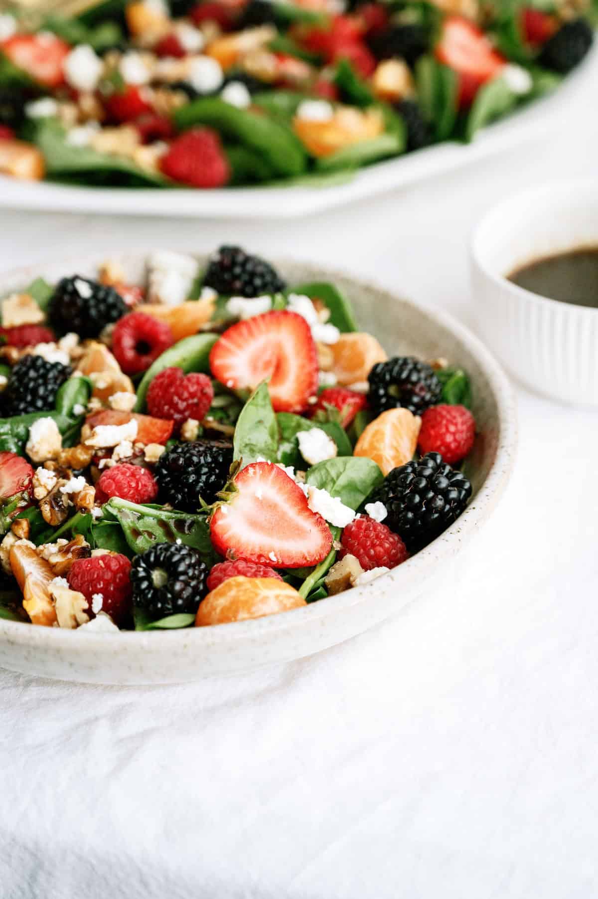 A bowl of fresh salad with spinach, strawberries, blackberries, raspberries, orange segments, walnuts, and feta cheese sits on a white tablecloth next to a small bowl of dark dressing.