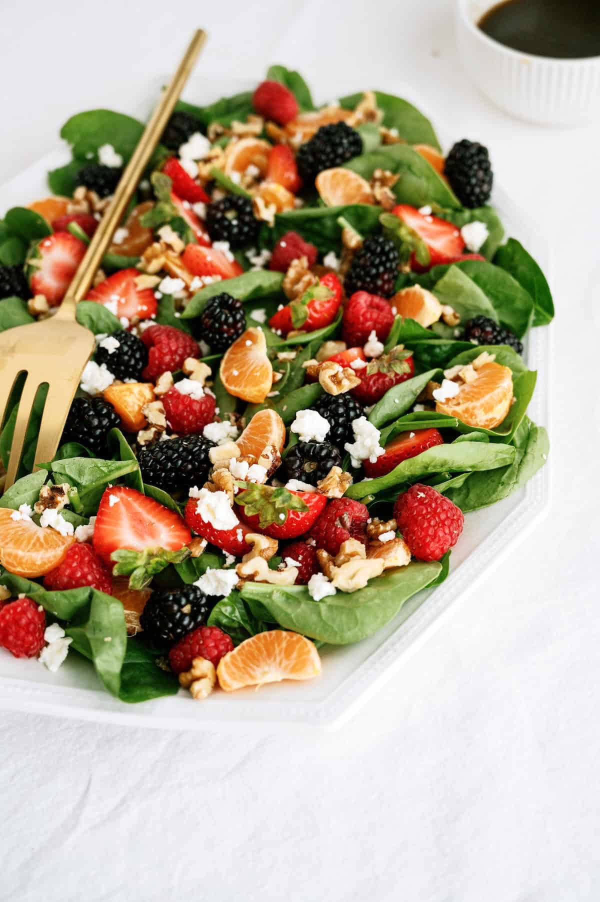 A plate of spinach salad topped with blackberries, raspberries, strawberries, mandarin orange segments, walnuts, and crumbled cheese, with a serving utensil on the side.