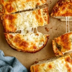 Sliced cheesy garlic bread on parchment paper, with melted cheese and herbs on top, next to a blue cloth.