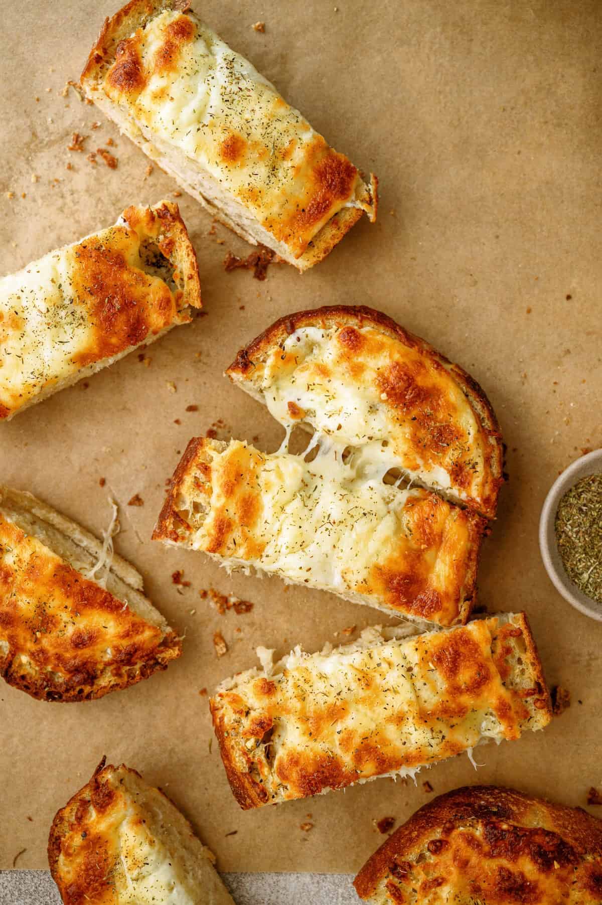 Slices of toasted garlic bread topped with melted cheese are arranged on parchment paper, with a small bowl of dried herbs on the side.