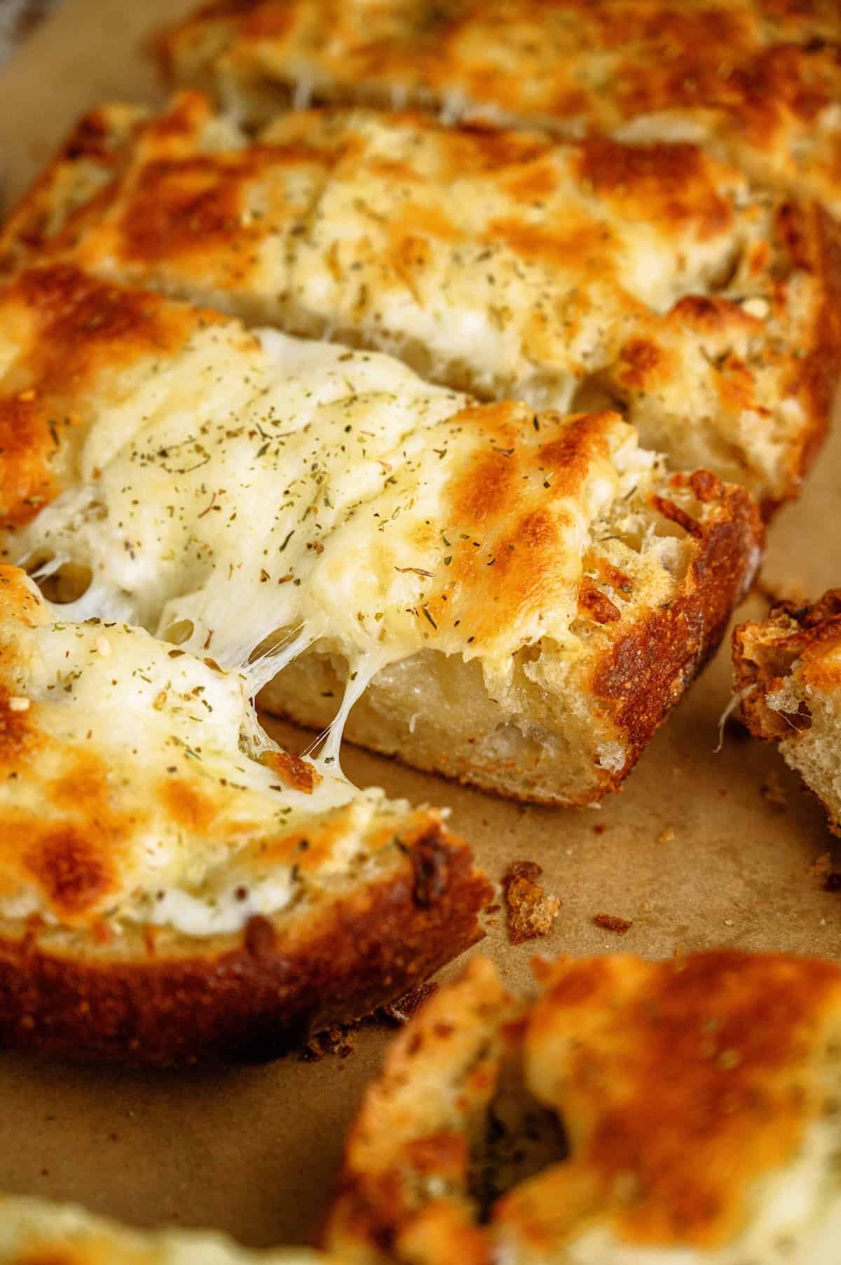 Slices of cheesy garlic bread with melted cheese and herbs on top, resting on a piece of brown parchment paper.