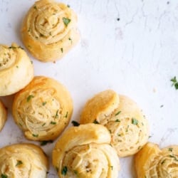 Seven golden brown, spiral-shaped bread rolls topped with chopped herbs are arranged on a white, lightly textured surface.