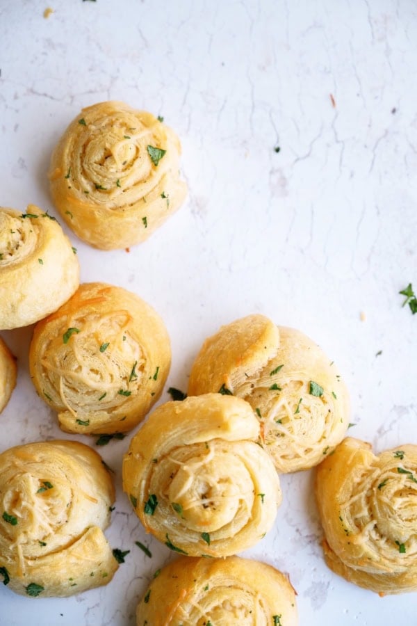 Seven golden brown, spiral-shaped bread rolls topped with chopped herbs are arranged on a white, lightly textured surface.