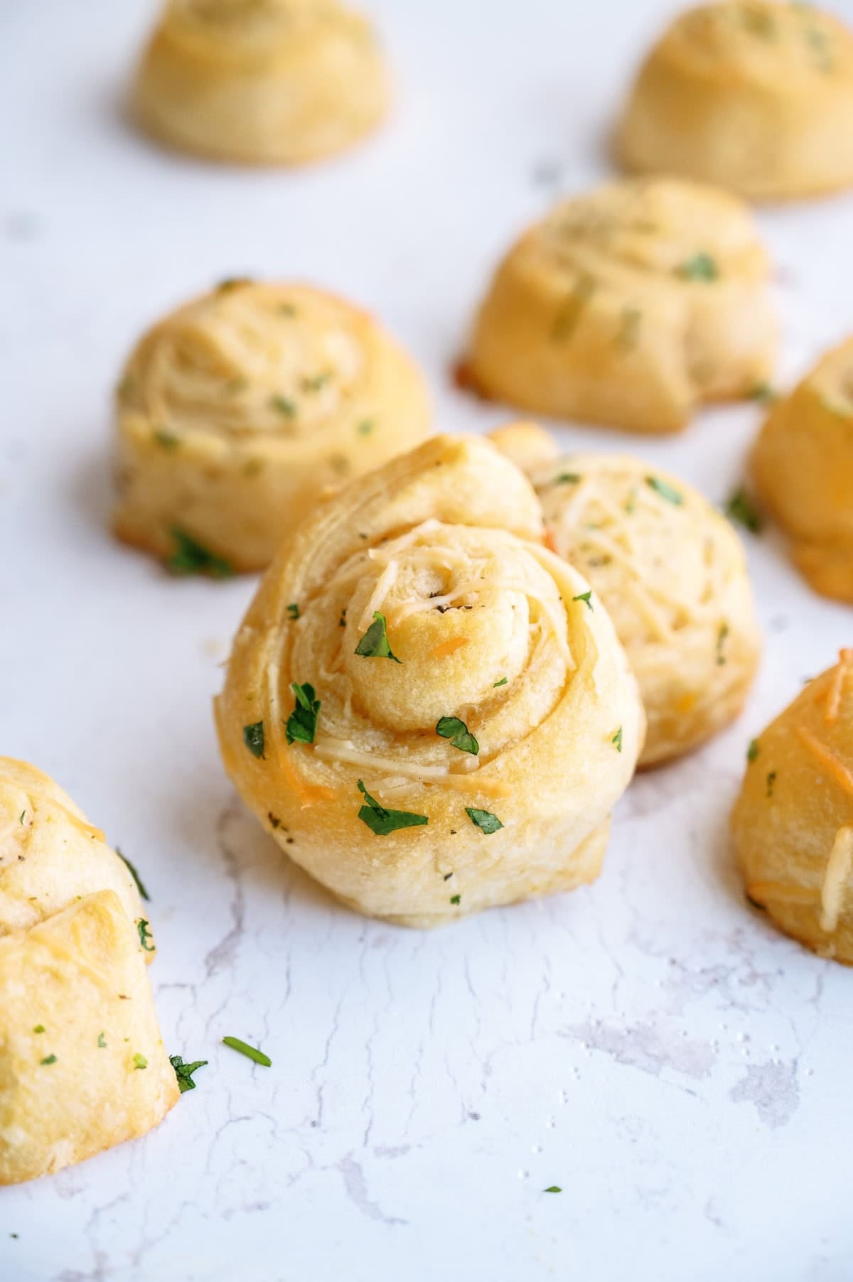 Several golden-brown garlic bread rolls garnished with chopped parsley are arranged on a white marble surface.