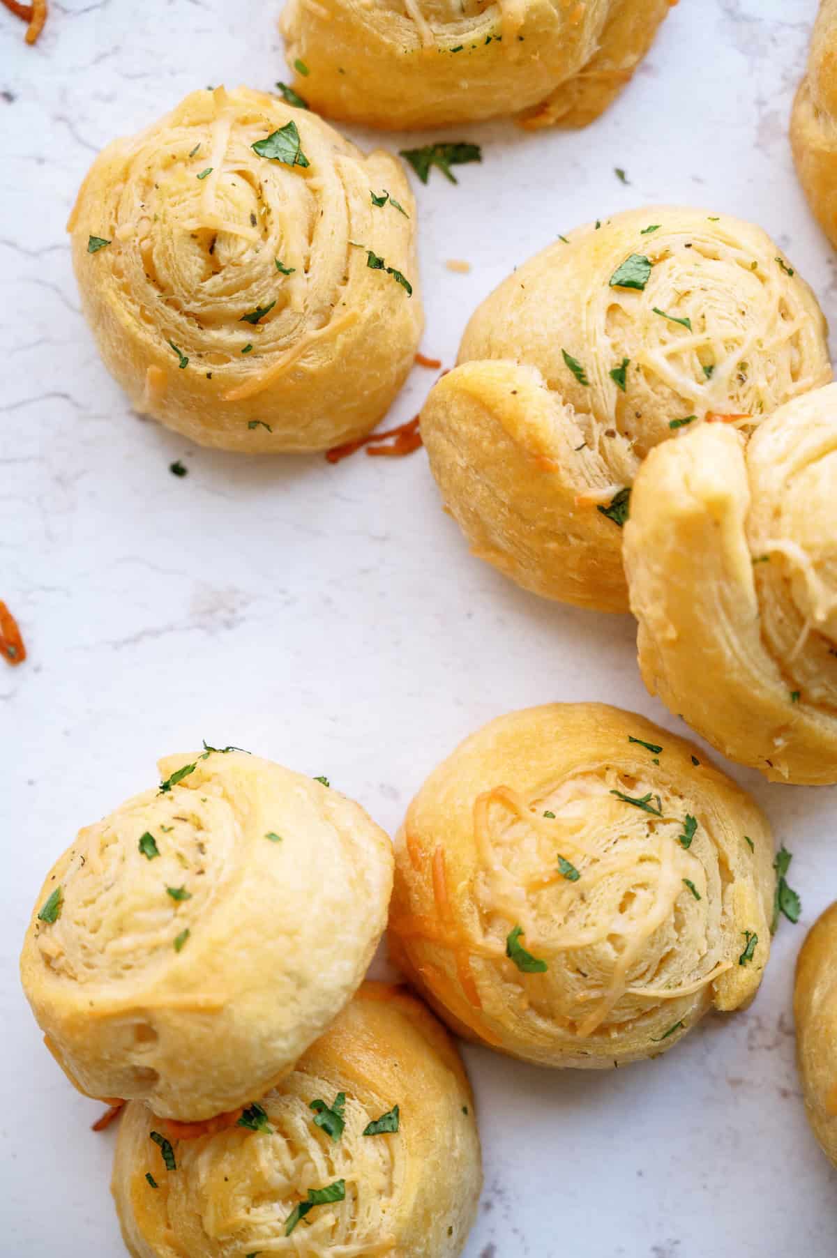 Golden brown, spiral-shaped bread rolls with shredded cheese and chopped herbs on a white surface.