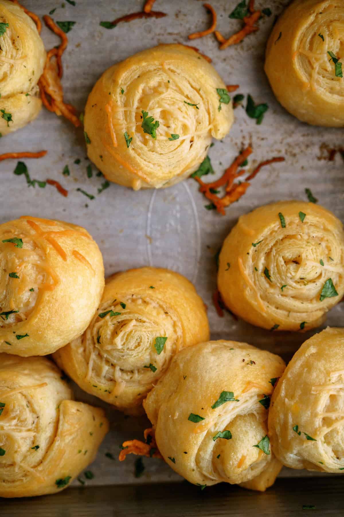 Baked garlic cheese rolls on a parchment-lined baking sheet, sprinkled with chopped parsley and shredded cheese.
