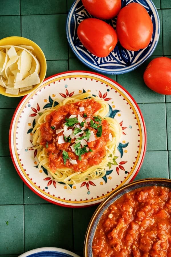 A plate of spaghetti topped with tomato sauce and parsley, surrounded by fresh tomatoes, a bowl of grated cheese, and extra sauce on a green tiled surface.