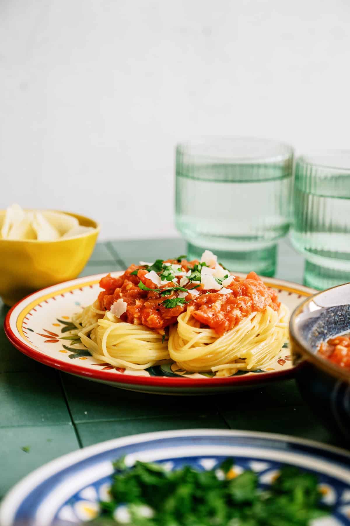 A plate of spaghetti topped with tomato sauce, parmesan cheese, and herbs sits on a green table next to two glasses of water and a small bowl of cheese.