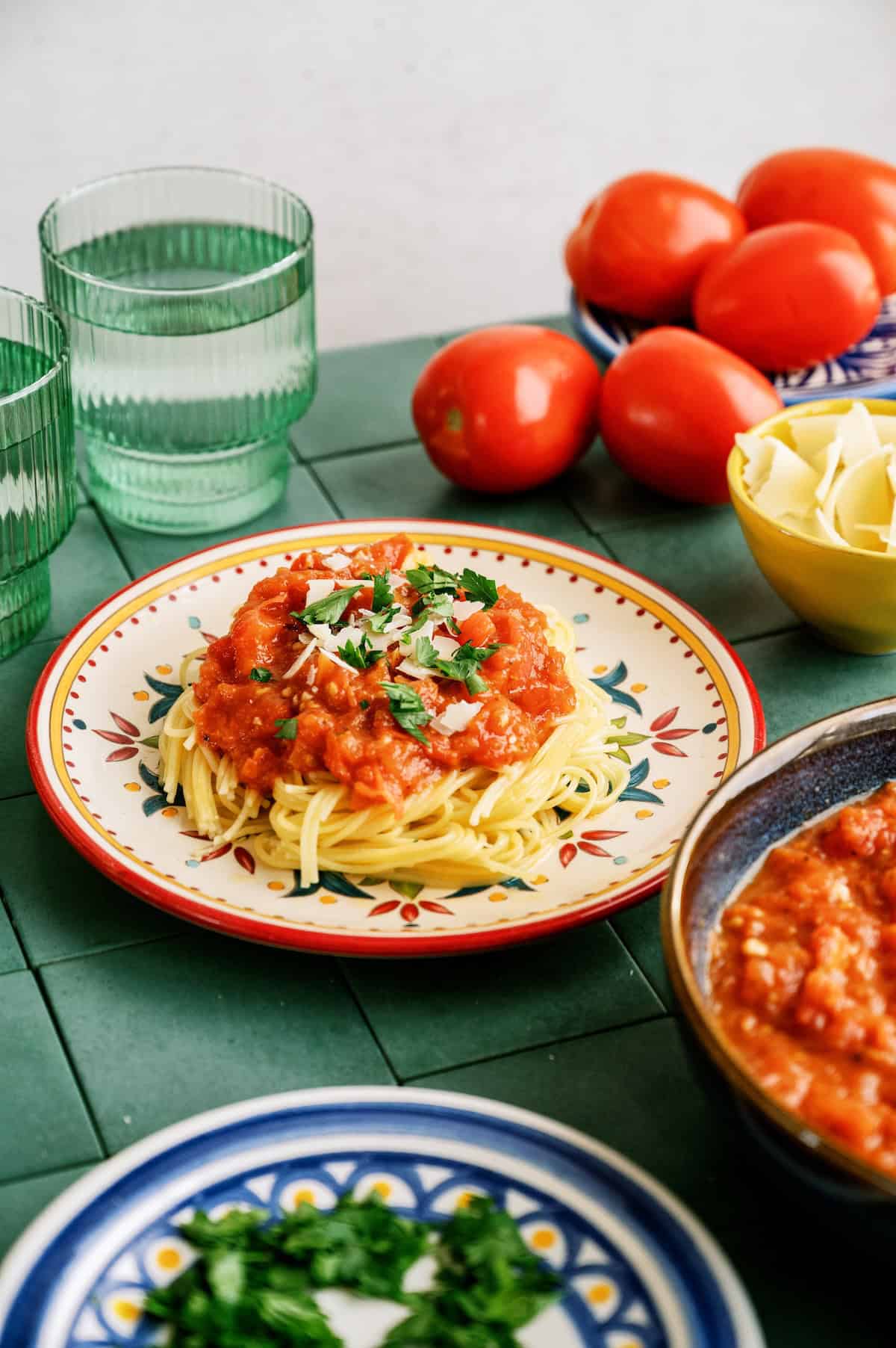 A plate of spaghetti topped with tomato sauce and herbs sits on a green tiled table, surrounded by tomatoes, a bowl of pasta ribbons, glassware, and a dish of chopped herbs.