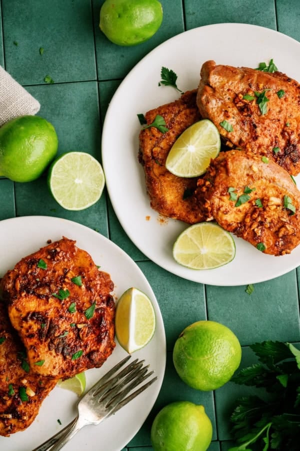 Two plates of seasoned, cooked chicken garnished with chopped herbs and lime wedges on a green tiled surface, surrounded by whole and halved limes.