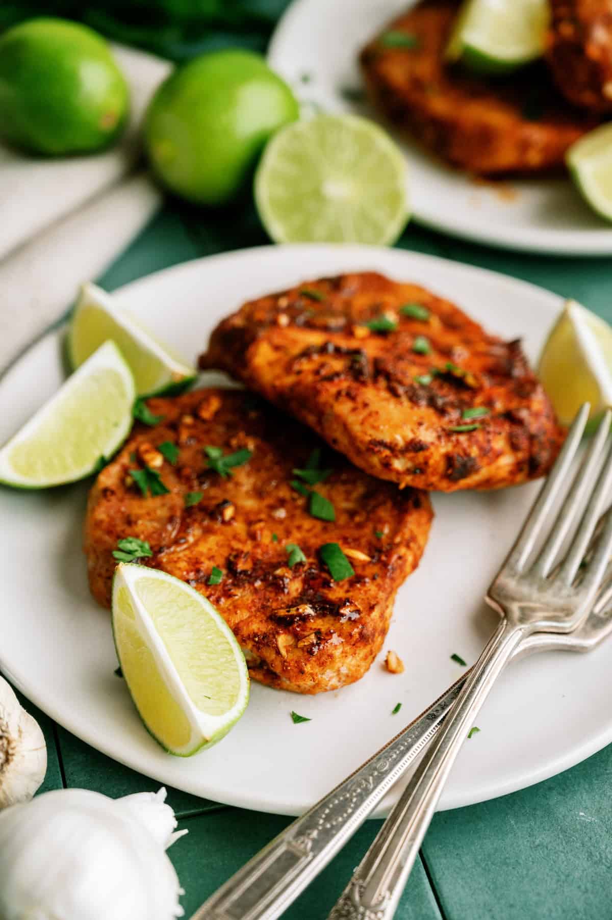 Two seasoned, cooked chicken breasts garnished with chopped herbs and lime wedges are arranged on a white plate with two metal forks.