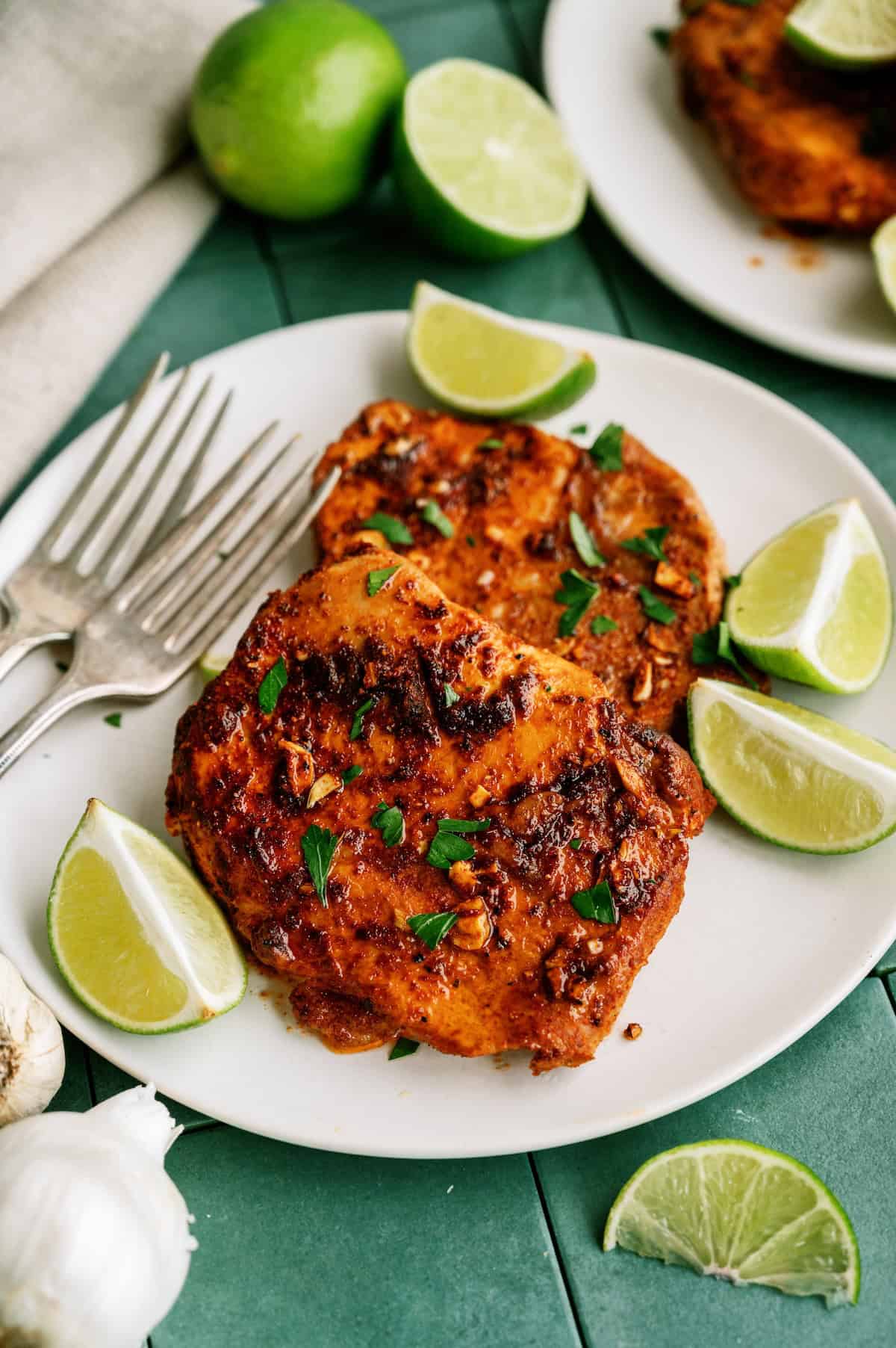A plate with two pieces of seasoned, grilled chicken garnished with chopped herbs and lime wedges, with two forks on the side. Garlic and whole limes are nearby.