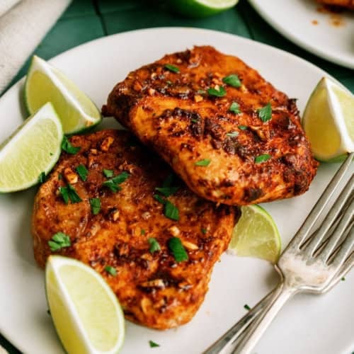 Two pieces of seasoned grilled chicken on a white plate, garnished with chopped herbs and lime wedges, with a fork and knife beside them.