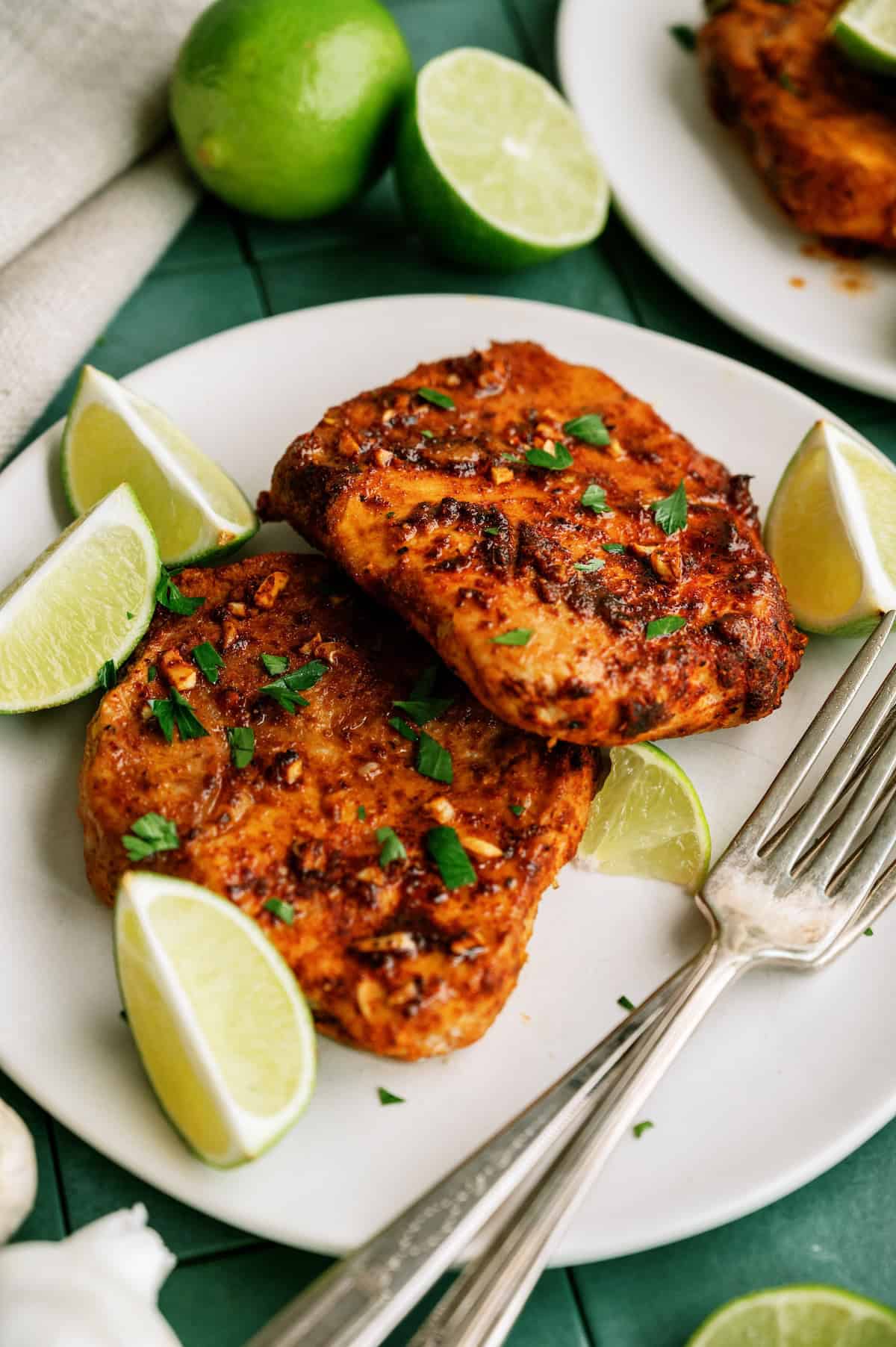 Two pieces of seasoned grilled chicken on a white plate, garnished with chopped herbs and lime wedges, with a fork and knife beside them.