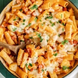 A pan of baked rigatoni pasta with tomato sauce, melted cheese, chopped herbs, and a wooden spoon on a green tiled surface.