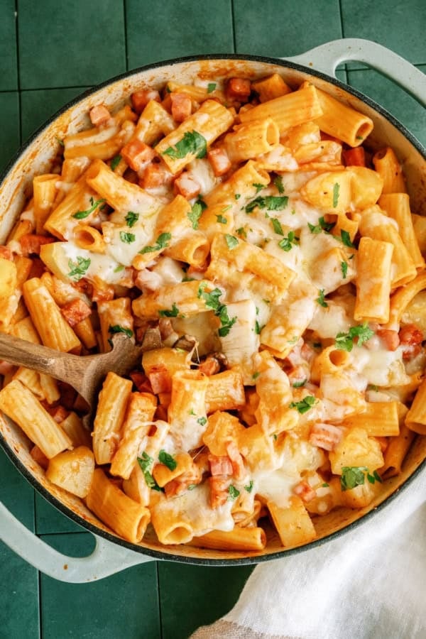 A pan of baked rigatoni pasta with tomato sauce, melted cheese, chopped herbs, and a wooden spoon on a green tiled surface.