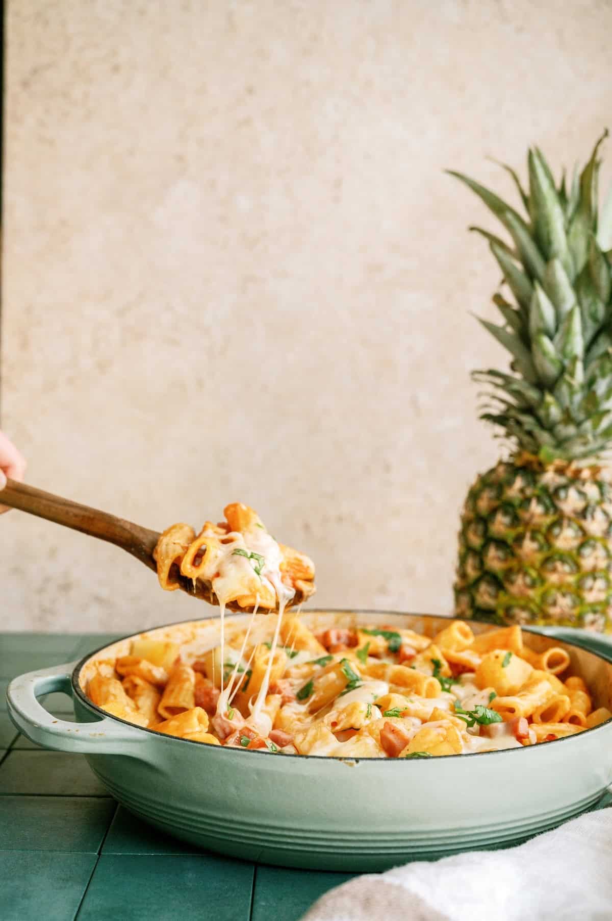 A green baking dish filled with cheesy baked pasta, with a wooden spoon lifting a serving; a whole pineapple sits in the background.