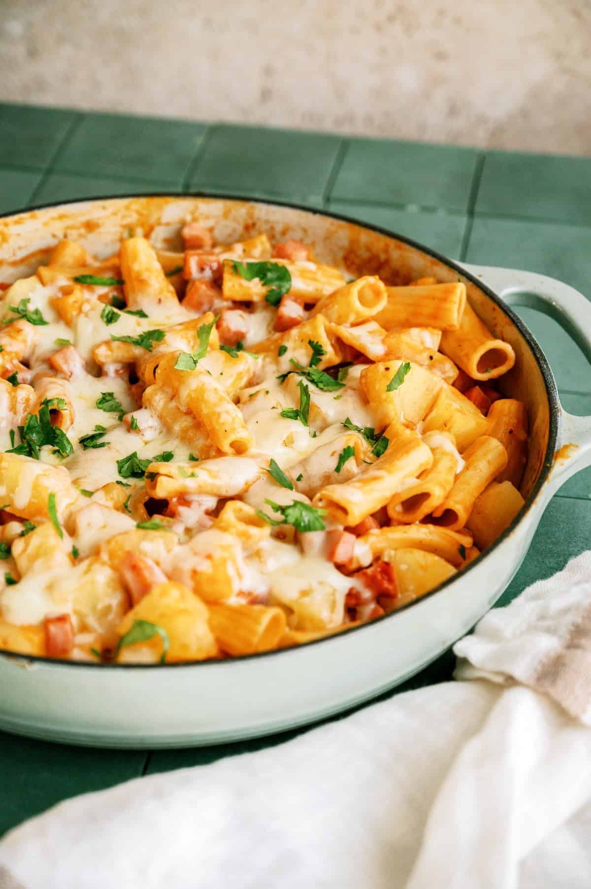 A skillet filled with baked rigatoni pasta, melted cheese, tomato sauce, and chopped parsley, placed on a green surface with a white cloth beside it.