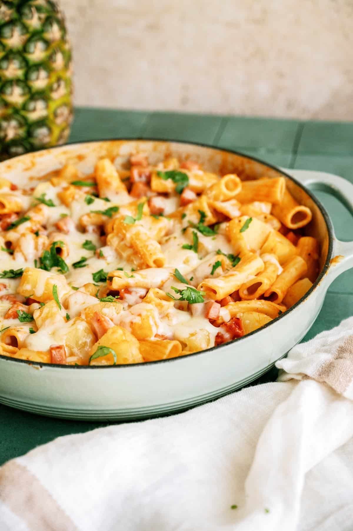 Baked pasta with cheese, ham, pineapple, and herbs is served in a round, green casserole dish on a green table next to a white cloth.