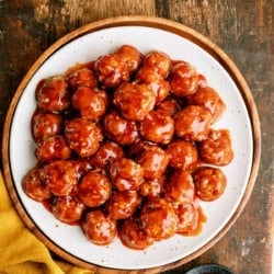 A plate of glazed meatballs arranged in a circular pattern, served with a small bowl of dipping sauce on a wooden table with a yellow cloth.