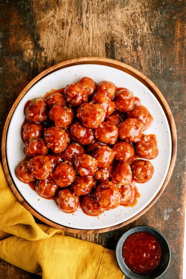 A plate of glazed meatballs arranged in a circular pattern, served with a small bowl of dipping sauce on a wooden table with a yellow cloth.