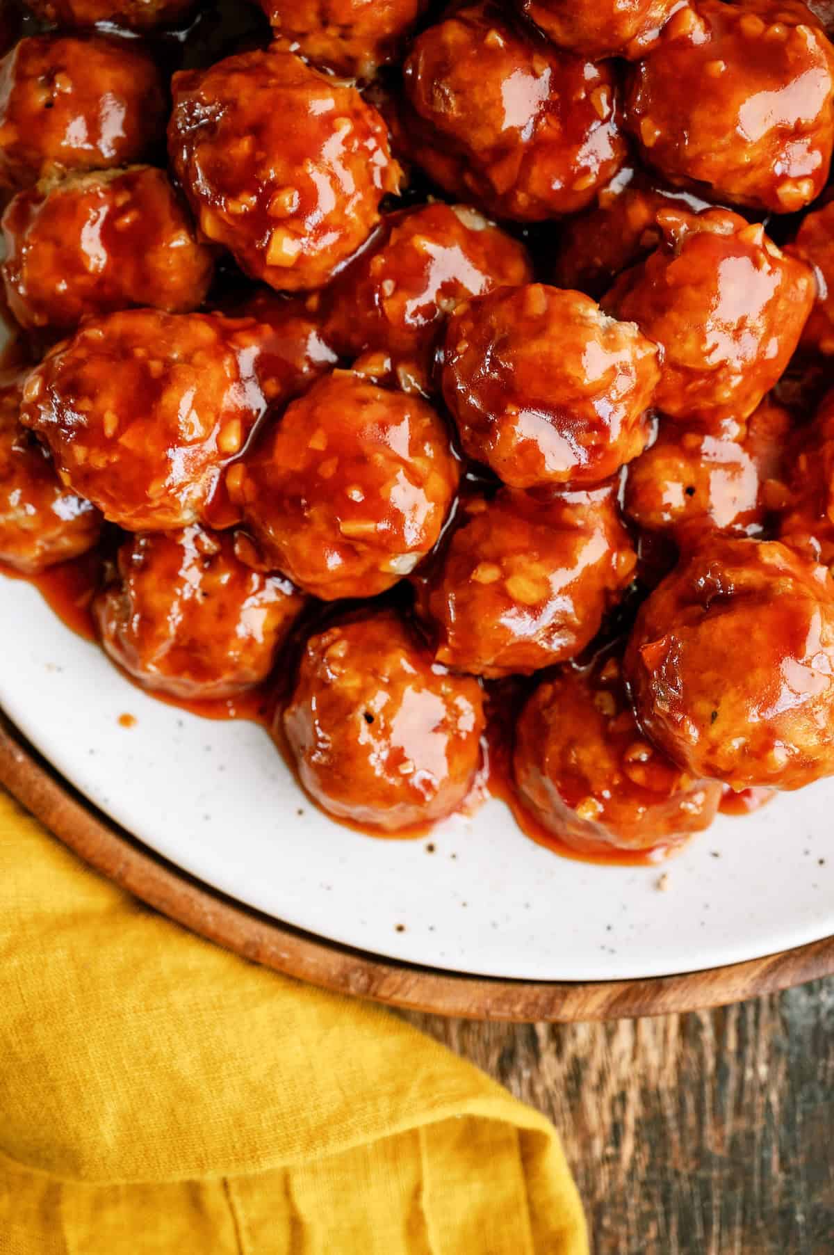 A close-up of saucy meatballs piled on a white plate, with a yellow cloth napkin nearby on a wooden surface.
