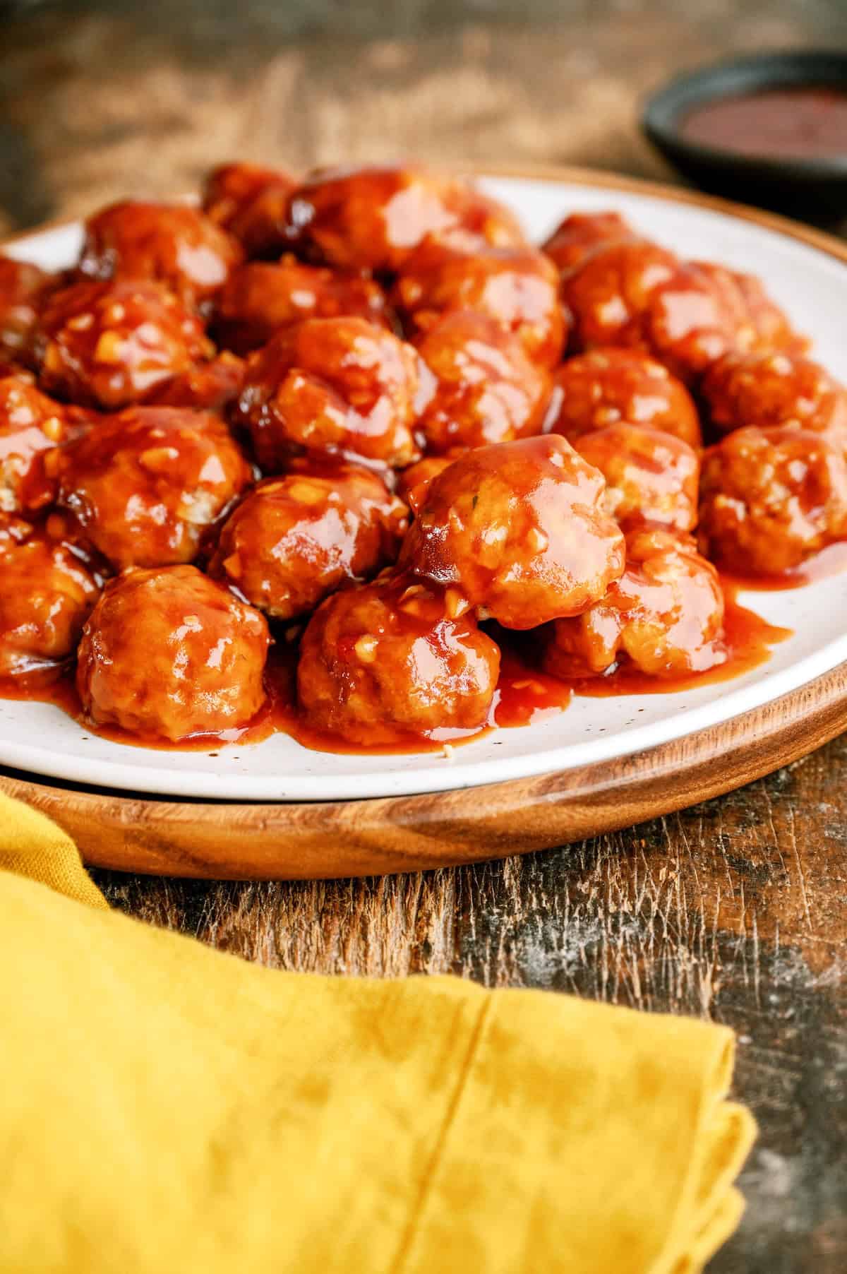 A white plate filled with glazed meatballs covered in sauce, set on a wooden surface with a yellow cloth nearby.