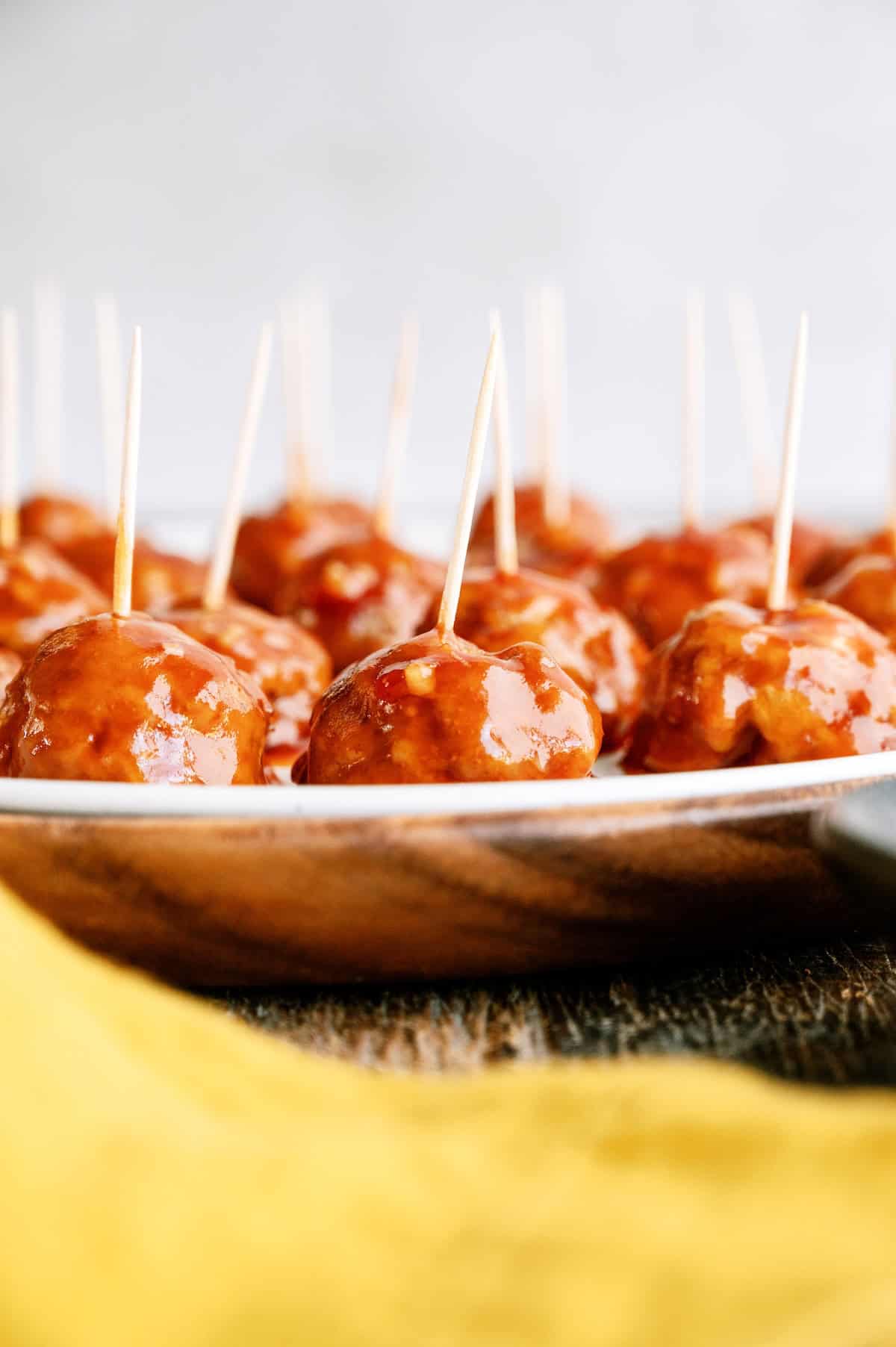 A plate of glazed meatballs, each with a toothpick inserted, displayed on a wooden surface with a yellow cloth in the foreground.