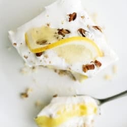 A slice of creamy lemon dessert topped with whipped cream, lemon slices, and chopped nuts on a white plate with a spoonful beside it.