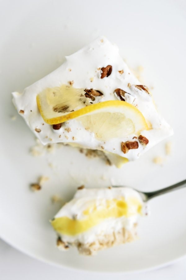A slice of creamy lemon dessert topped with whipped cream, lemon slices, and chopped nuts on a white plate with a spoonful beside it.