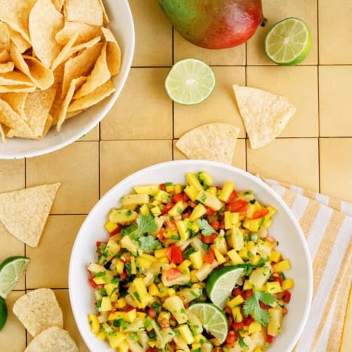 A bowl of mango salsa with cilantro and lime, surrounded by tortilla chips, whole mangoes, and lime halves on a tiled surface.