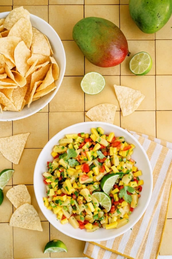 A bowl of mango salsa with cilantro and lime, surrounded by tortilla chips, whole mangoes, and lime halves on a tiled surface.