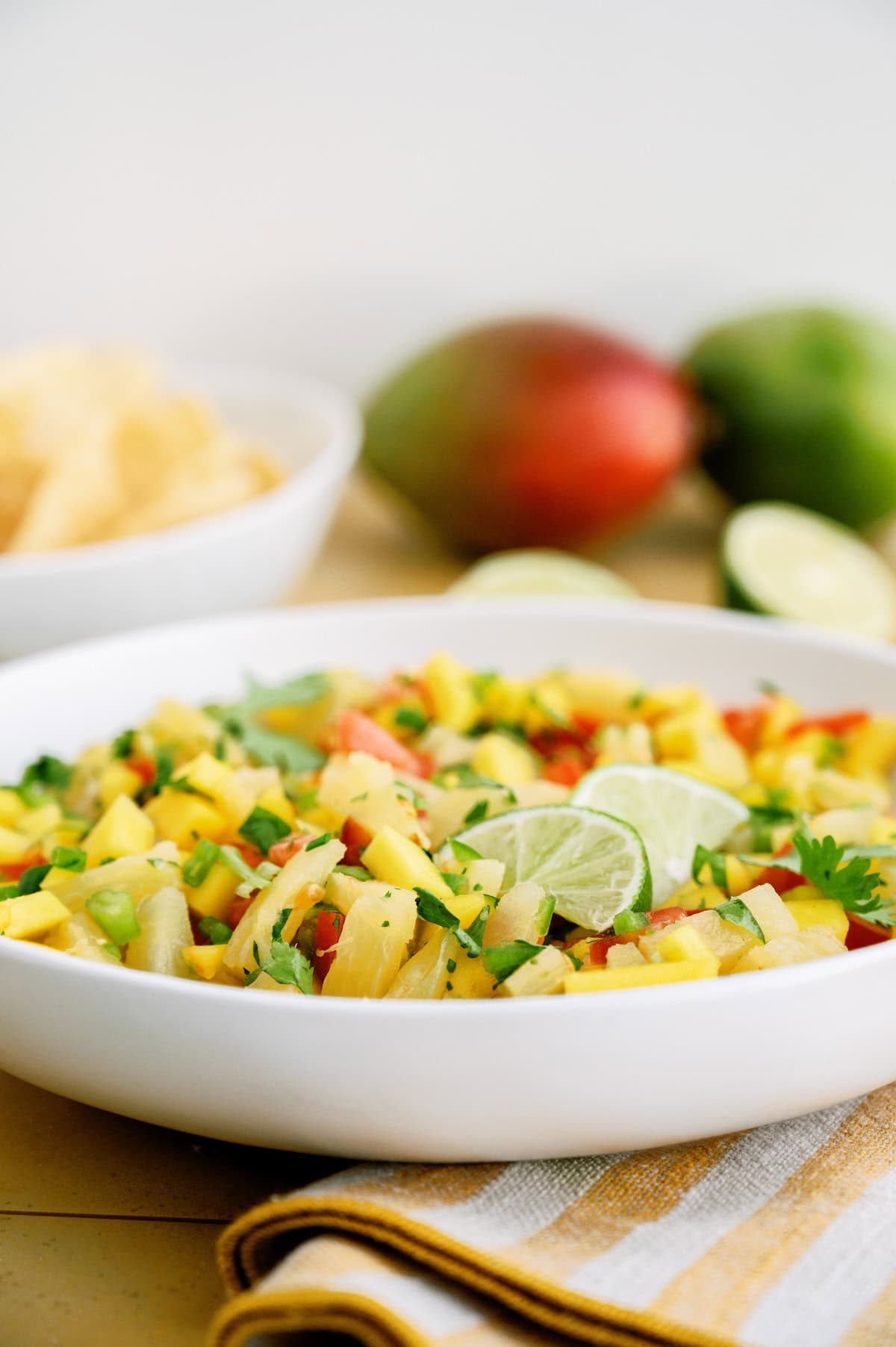 A white bowl filled with mango salsa garnished with lime slices sits on a table, with tortilla chips and whole mangoes in the background.