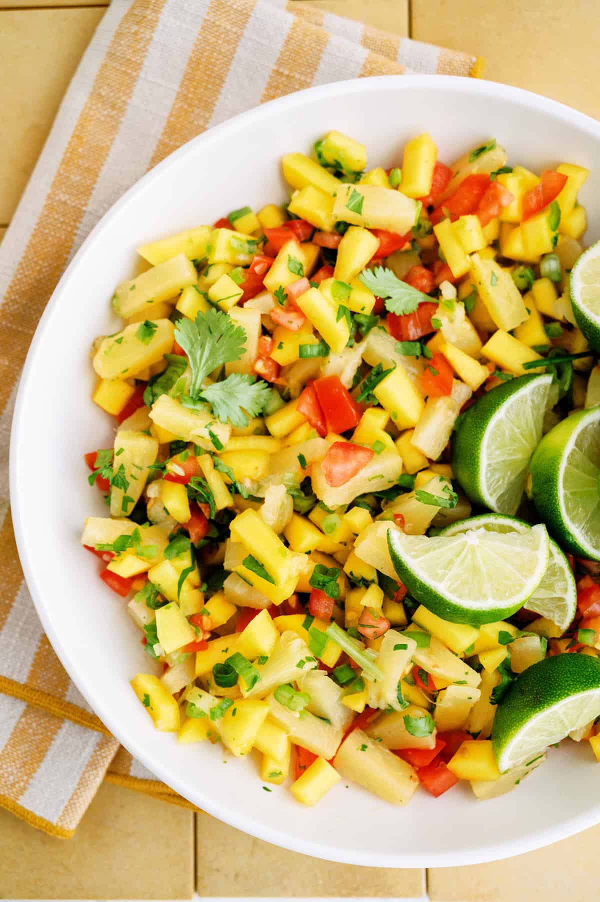 A bowl of chopped mango salsa with red bell peppers, onions, cilantro, and lime wedges, placed on a striped cloth.