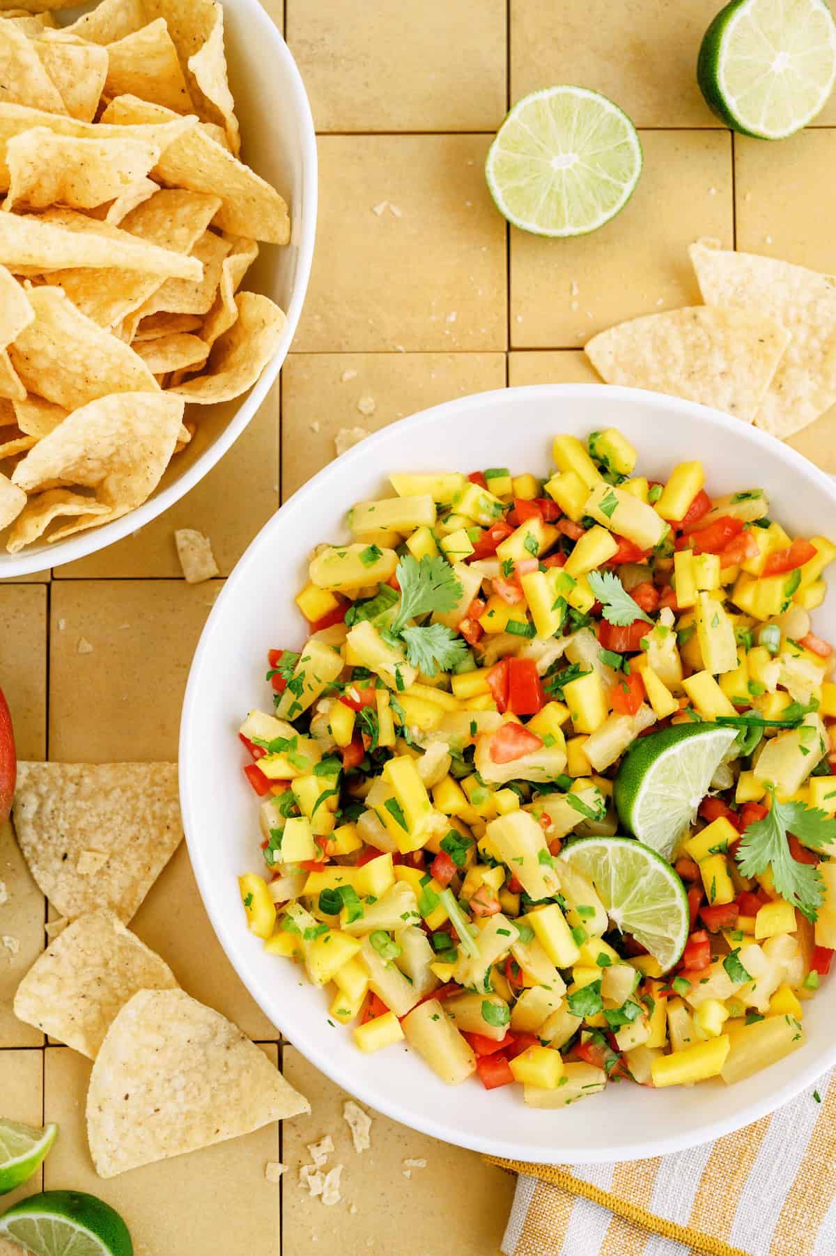 A bowl of mango salsa garnished with lime and cilantro sits next to a bowl of tortilla chips on a tiled surface.