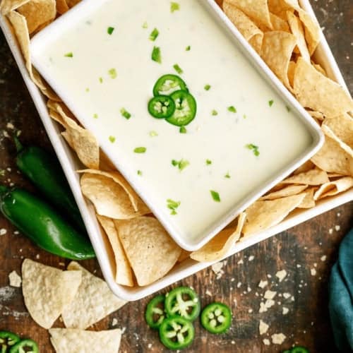 A rectangular dish of white cheese dip garnished with sliced jalapeños, surrounded by tortilla chips, on a rustic wooden surface with scattered jalapeño slices and chips.