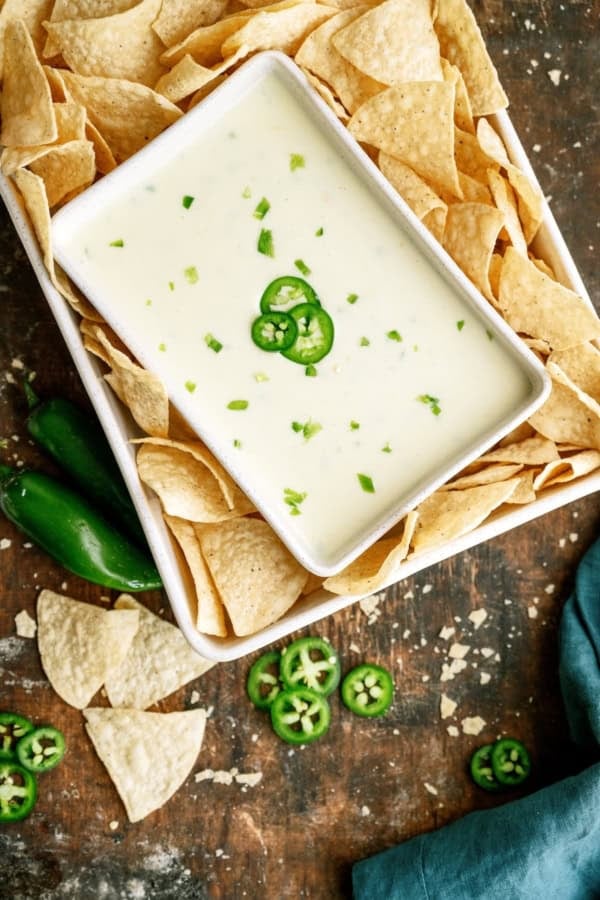 A rectangular dish of white cheese dip garnished with sliced jalapeños, surrounded by tortilla chips, on a rustic wooden surface with scattered jalapeño slices and chips.