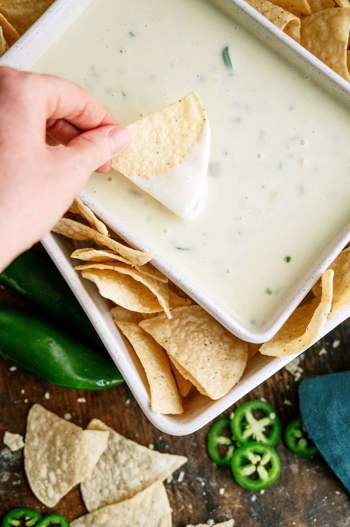 A hand dips a tortilla chip into a rectangular dish of white cheese queso dip, surrounded by more chips and sliced jalapeños.