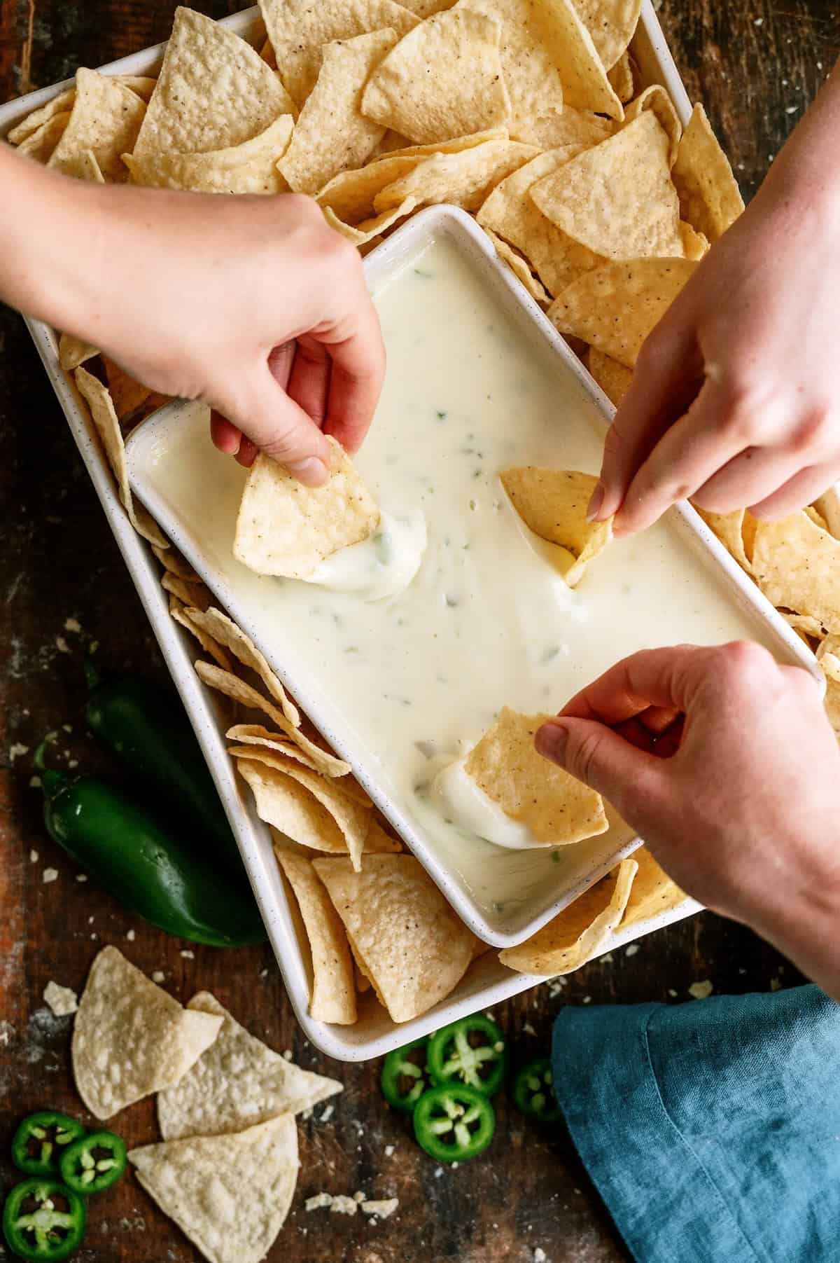 Three hands dip tortilla chips into a tray of white cheese queso, surrounded by more chips on a rustic wooden table with jalapeño slices nearby.