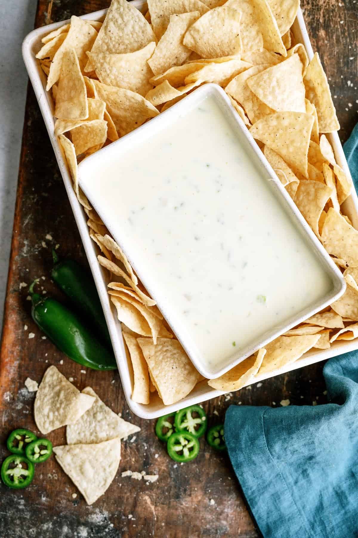 A rectangular tray filled with tortilla chips surrounds a dish of white queso dip; sliced and whole jalapeños are on the wooden surface nearby.