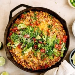 A cast iron skillet filled with a colorful Mexican-inspired casserole topped with melted cheese, tomatoes, lettuce, cilantro, and lime slices, surrounded by lime wedges and herbs.