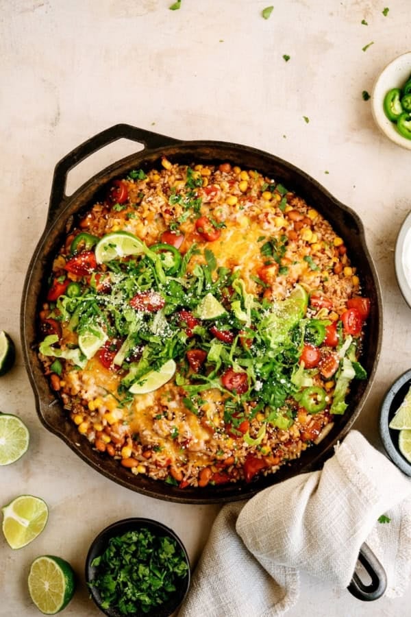 A cast iron skillet filled with a colorful Mexican-inspired casserole topped with melted cheese, tomatoes, lettuce, cilantro, and lime slices, surrounded by lime wedges and herbs.