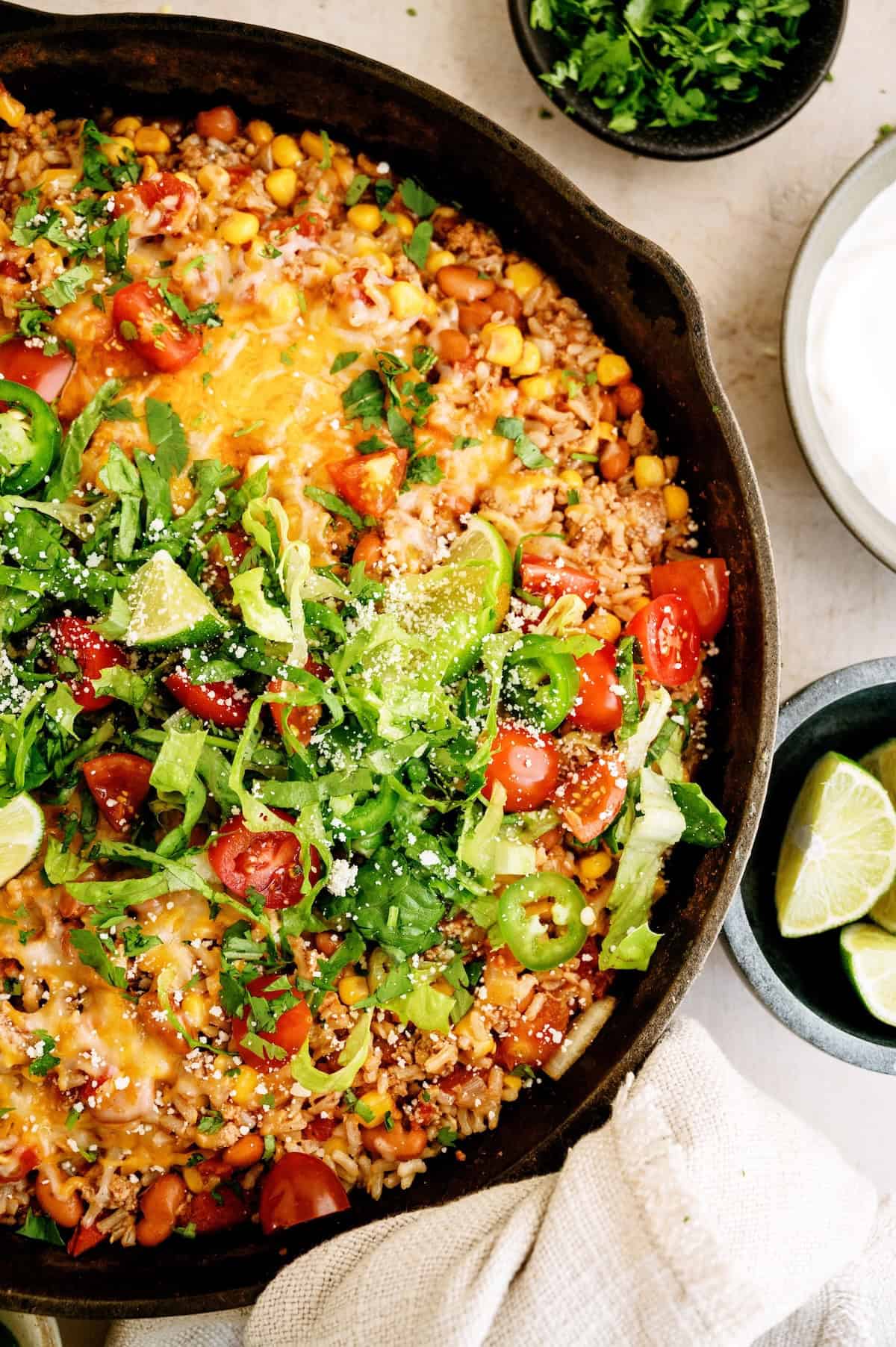 A cast iron skillet filled with Mexican-inspired rice, beans, corn, melted cheese, lettuce, tomatoes, jalapeños, lime wedges, and garnished with cilantro. Sour cream and extra limes on the side.